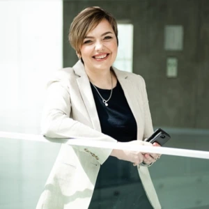 Happy woman with short light-brown hair, wearing a beige blazer over a black top and holding a smartphone while leaning against a glass railing in a bright modern interior.