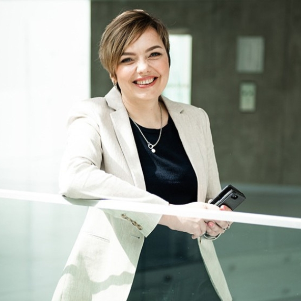Happy woman with short light-brown hair, wearing a beige blazer over a black top and holding a smartphone while leaning against a glass railing in a bright modern interior.