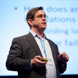 A man wearing glasses, a suit jacket, and a light-colored shirt while speaking at an event. He is shown in profile against a blurred presentation background.