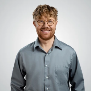 Happy man with curly light-brown hair, round glasses and a short beard, wearing a grey button-up shirt against a light studio background.