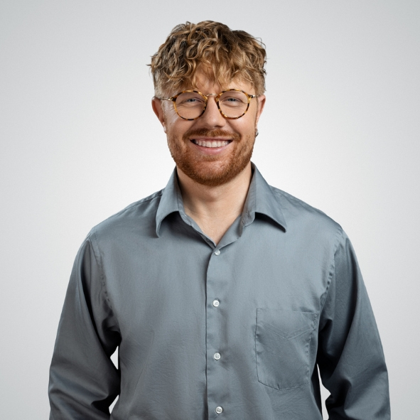 Happy man with curly light-brown hair, round glasses and a short beard, wearing a grey button-up shirt against a light studio background.
