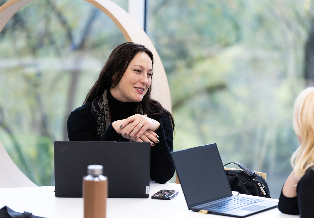 Woman in a black turtleneck speaking across a table with two open laptops during an Open Innovation Centre forum discussion.