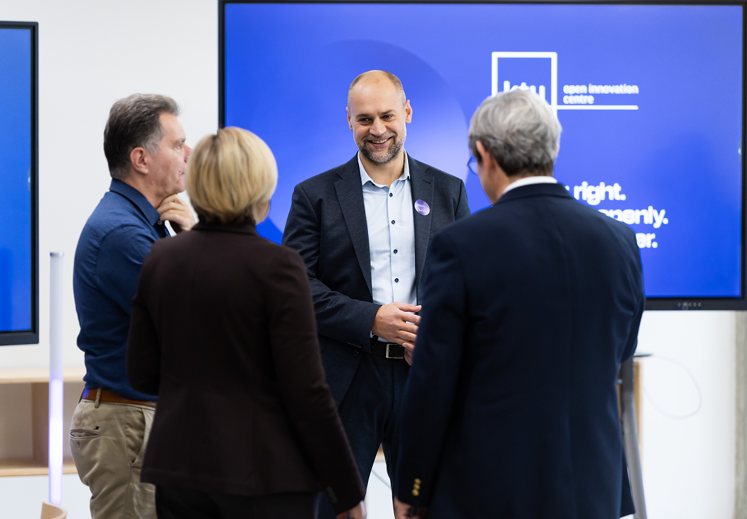 Four professionals standing in conversation during a networking session at the Open Innovation Centre forum with an innovation presentation displayed behind them.