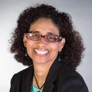 Portrait of a smiling woman with curly dark hair and red glasses, wearing a black blazer and turquoise top against a light grey background.