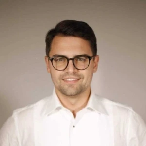 Portrait of a smiling man with short dark hair and round glasses, wearing a white shirt against a neutral beige background.