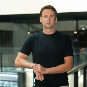 Portrait of a man in a black T-shirt leaning on a glass railing in a modern indoor setting.