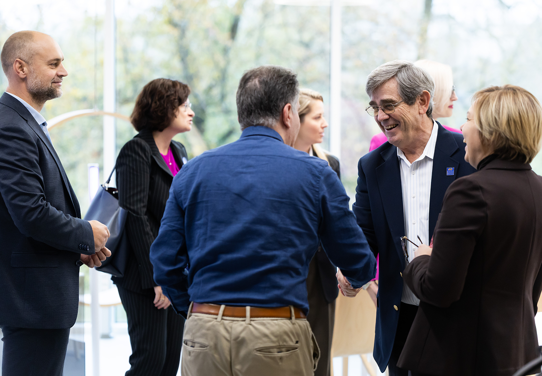 OIC for Follow-on collaboration opportunities Group of professionals networking and conversing in a bright modern event space, with two men greeting each other in the foreground.