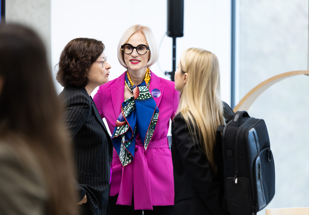 OIC for Open innovation community exposure Two women talking at a professional event, one wearing a bright pink suit, patterned scarf and black glasses, and the other dressed in a dark pinstripe suit.
