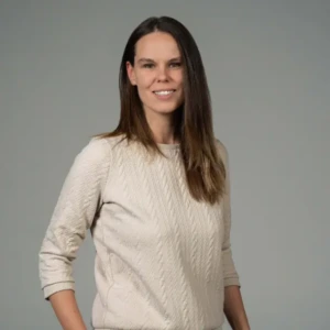 Portrait of a smiling woman with long brown hair, wearing a light beige cable-knit sweater, standing against a plain grey background.